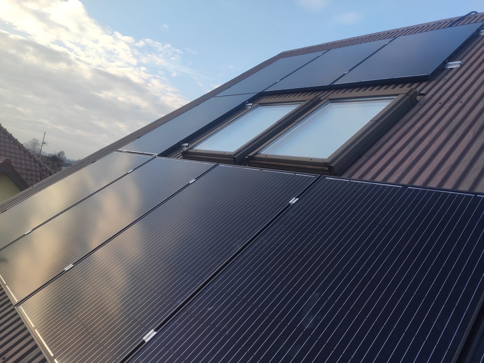Panele słoneczne zamontowane na dachu z dachówką, widoczne dwa okna dachowe, pochmurne niebo w tle.