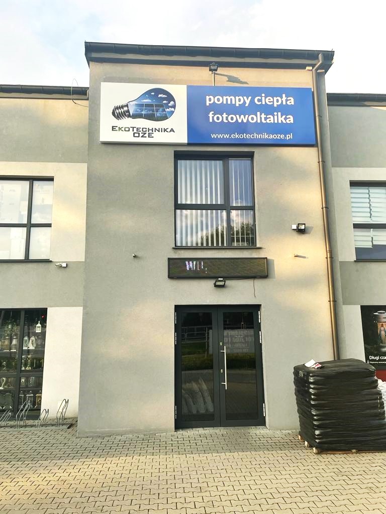 Fasada budynku firmy EkoTechnika OZE z reklamą pomp ciepła i fotowoltaiki, widoczne okna z żaluzjami, czarne drzwi wejściowe i paleta z workami pod ścianą.