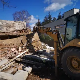 Żółta koparka CAT z łyżką na tle gruzów i resztek zburzonego budynku, błękitne niebo z chmurami w tle.