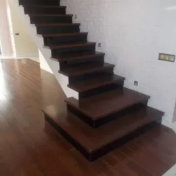 Nowoczesne, drewniane schody z ciemnymi stopniami i białymi podstopnicami, prowadzące w górę przy białej ścianie z fakturą cegły. Parkiet z ciemnego drewna na podłodze.