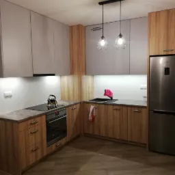 Nowoczesna kuchnia w odcieniach szarości i drewna z wiszącymi lampami w stylu industrialnym, czajnik na płycie indukcyjnej, lodówka w zabudowie.