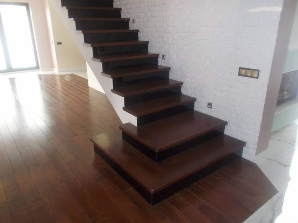 Nowoczesne, drewniane schody z ciemnymi stopniami i białymi podstopnicami, prowadzące w górę przy białej ścianie z fakturą cegły. Parkiet z ciemnego drewna na podłodze.