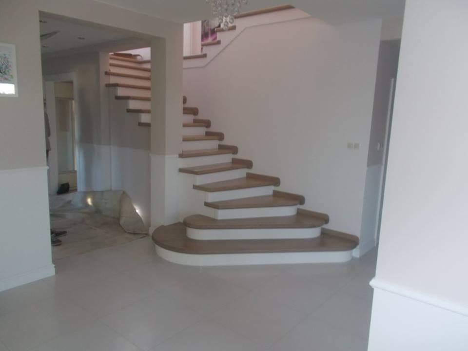 Drewniane schody z białymi podstopnicami, zakręcające w lewo, z widocznym fragmentem balustrady na górze i białymi płytkami na podłodze.