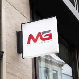 Podświetlany szyld z logo 'MG Mediagear' na elewacji budynku z beżowego kamienia, widoczna refleksja budynku w szybie okiennej.