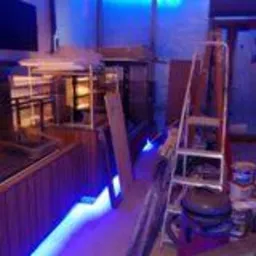 Wnętrze pomieszczenia w trakcie remontu, widoczne zabezpieczone folią ściany, drabina, puszki z farbą, elementy zabudowy barowej w trakcie montażu, oświetlenie LED w kolorze niebieskim.