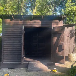 Nowa, ciemnobrązowa konstrukcja z bali z otwartymi drzwiami, prawdopodobnie składzik na narzędzia ogrodowe, z rampą wejściową i dwoma pilarkami łańcuchowymi na ziemi. W tle widoczne drzewa.