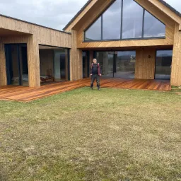 Nowoczesny dom szkieletowy z dużymi przeszkleniami i tarasem z drewna egzotycznego; mężczyzna w roboczym ubraniu stoi na tarasie, na trawniku leżą resztki materiałów budowlanych.