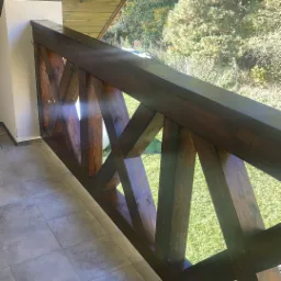 Kątowe ujęcie na ciemnobrązową, drewnianą balustradę z krzyżowym wzorem na balkonie z szarą podłogą. W tle zieleń drzew i trawnik w słoneczny dzień.