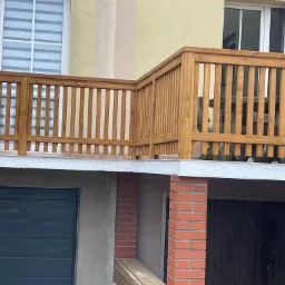 Drewniana balustrada tarasu z widocznymi pionowymi szczebelkami, jasnym wykończeniem i częściowo widocznym stołem z krzesłami, na tle elewacji budynku z oknami i dwoma garażami.