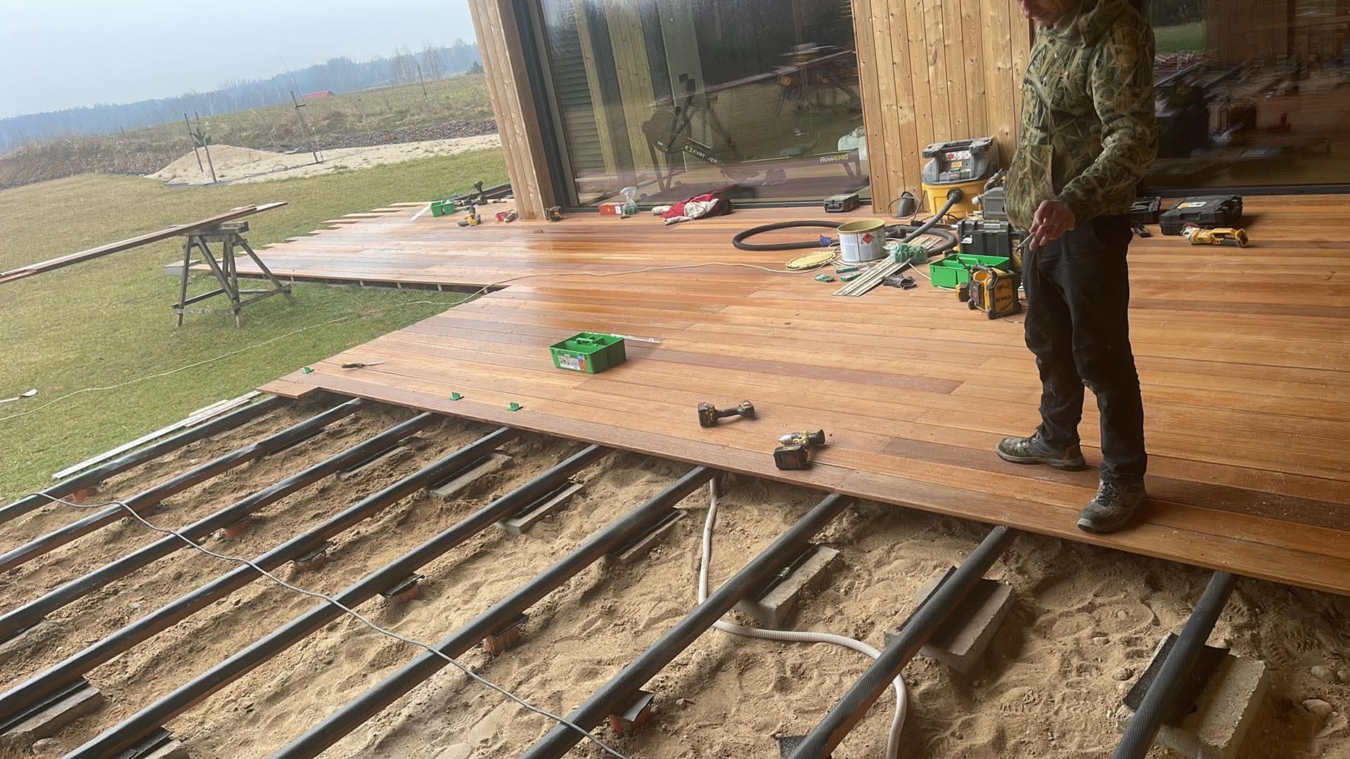 Montaż tarasu drewnianego z widoczną konstrukcją nośną i narzędziami; częściowo ułożone deski tarasowe w ciepłym odcieniu brązu.