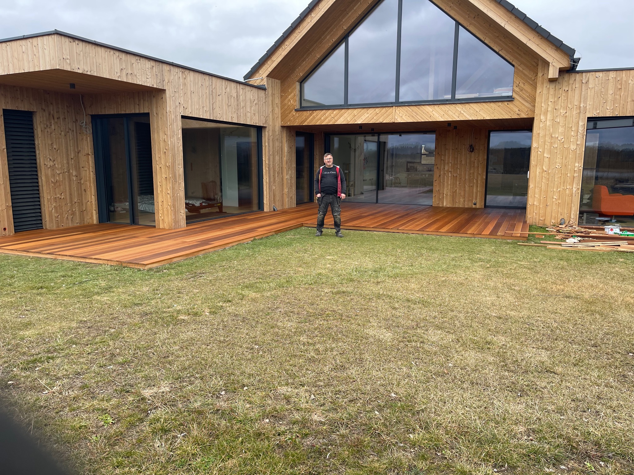 Nowoczesny dom szkieletowy z dużymi przeszkleniami i tarasem z drewna egzotycznego; mężczyzna w roboczym ubraniu stoi na tarasie, na trawniku leżą resztki materiałów budowlanych.