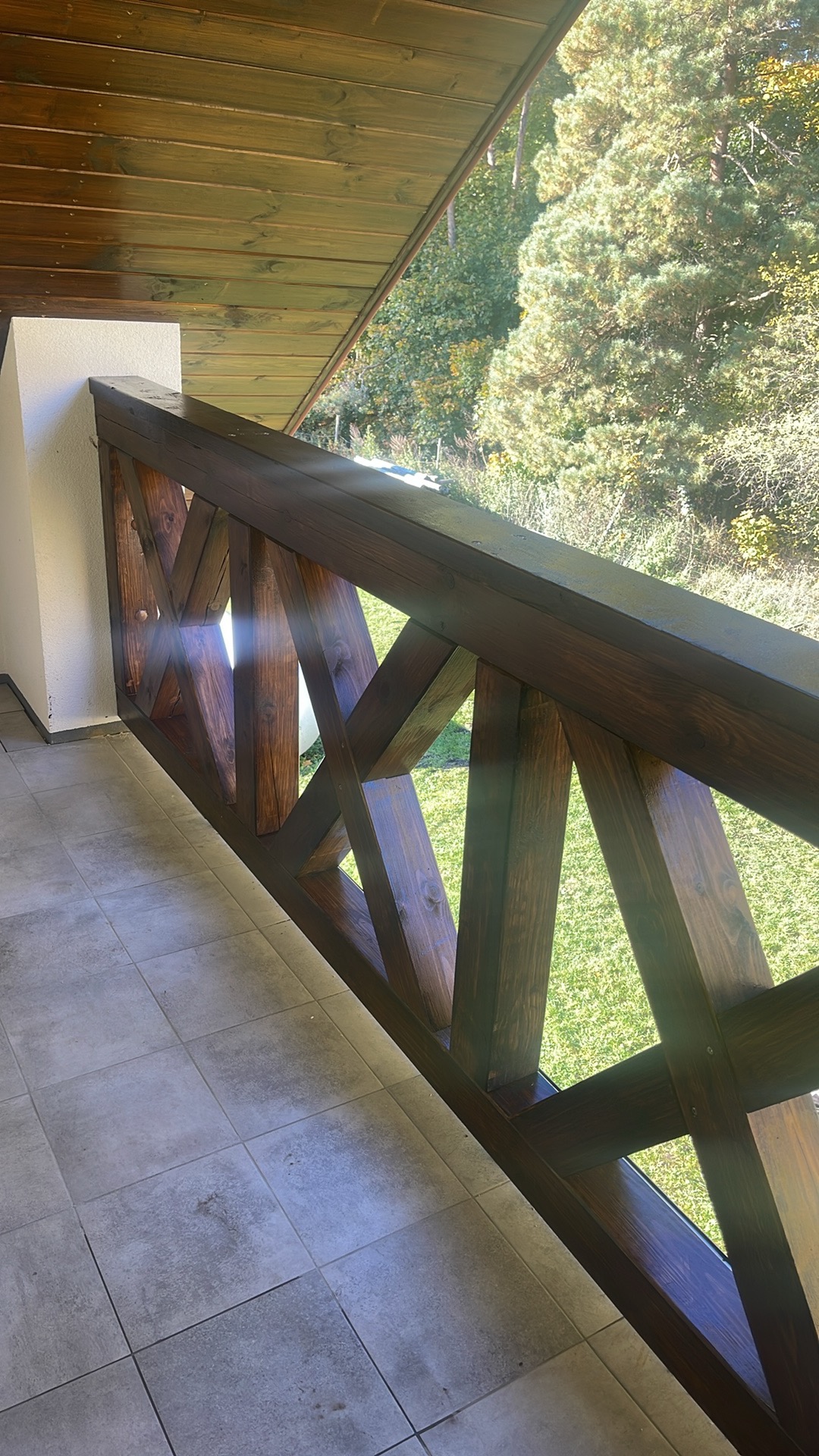 Kątowe ujęcie na ciemnobrązową, drewnianą balustradę z krzyżowym wzorem na balkonie z szarą podłogą. W tle zieleń drzew i trawnik w słoneczny dzień.