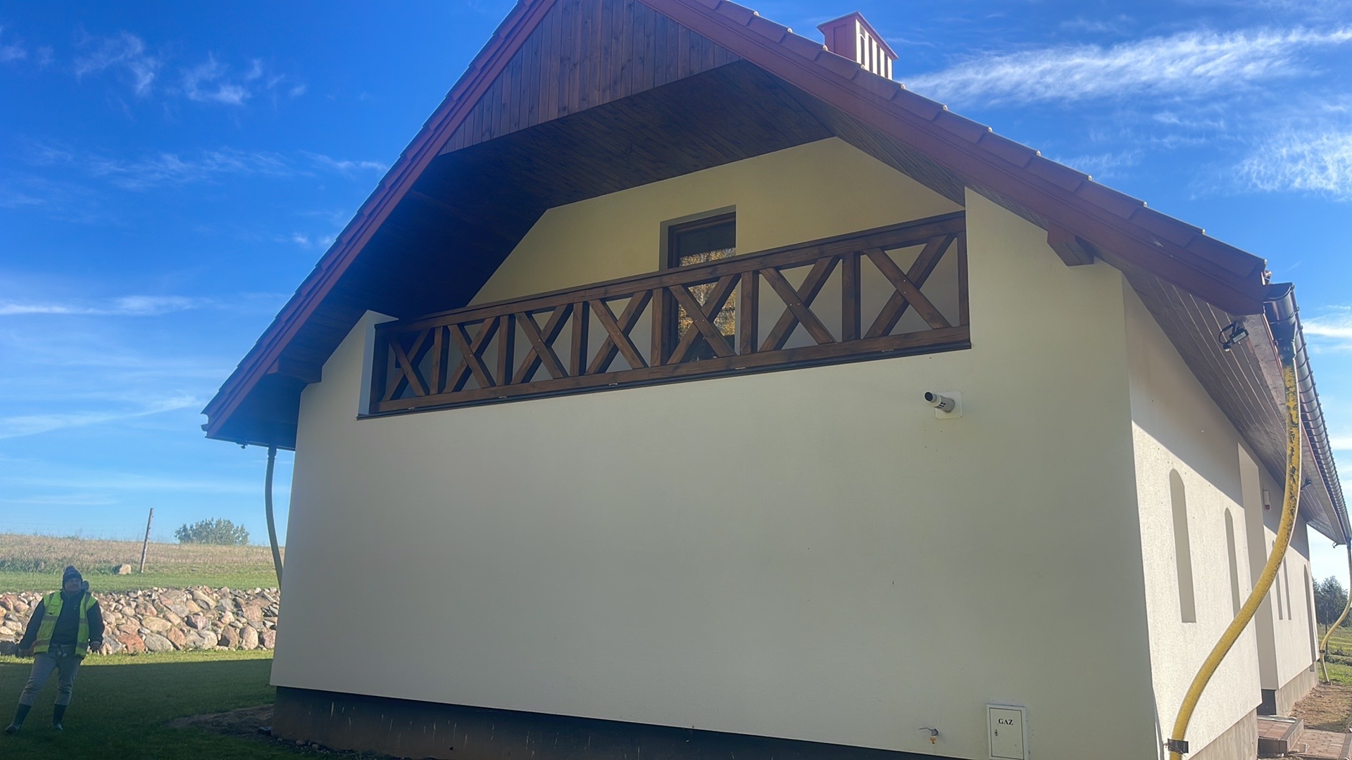 Boczny widok domu z beżową elewacją i drewnianą balustradą balkonu z motywem krzyży. Widoczny fragment dachu z brązową dachówką i osoba w oddali.