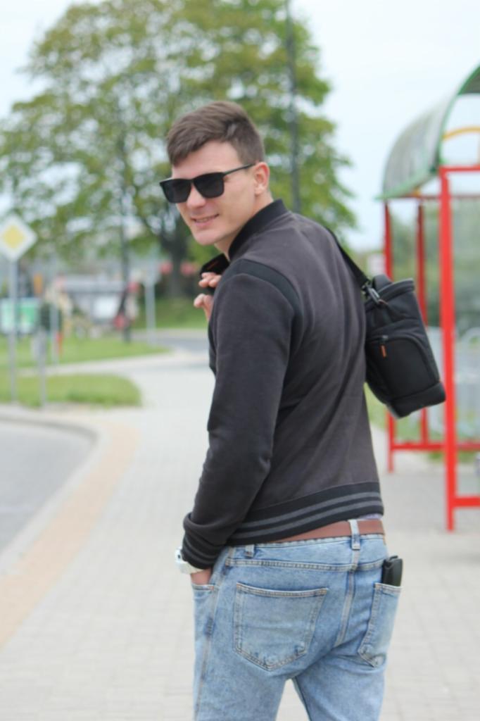 Mężczyzna w ciemnej kurtce i jeansach odwraca się z uśmiechem, na tle chodnika i zieleni. Na ramieniu ma czarną torbę. W tle czerwona konstrukcja przystanku.