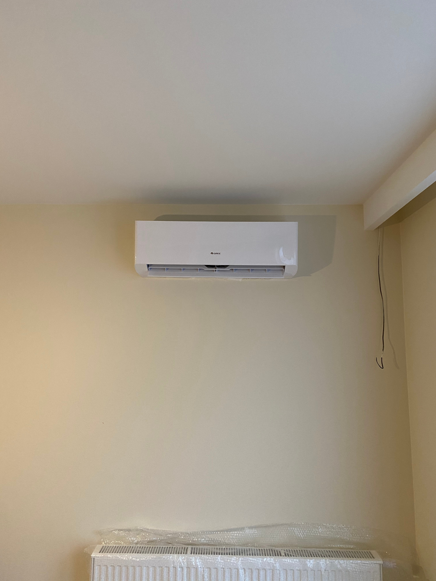 Biały klimatyzator marki Gree zamontowany na beżowej ścianie w pomieszczeniu, z widocznym grzejnikiem zabezpieczonym folią na dole kadru.