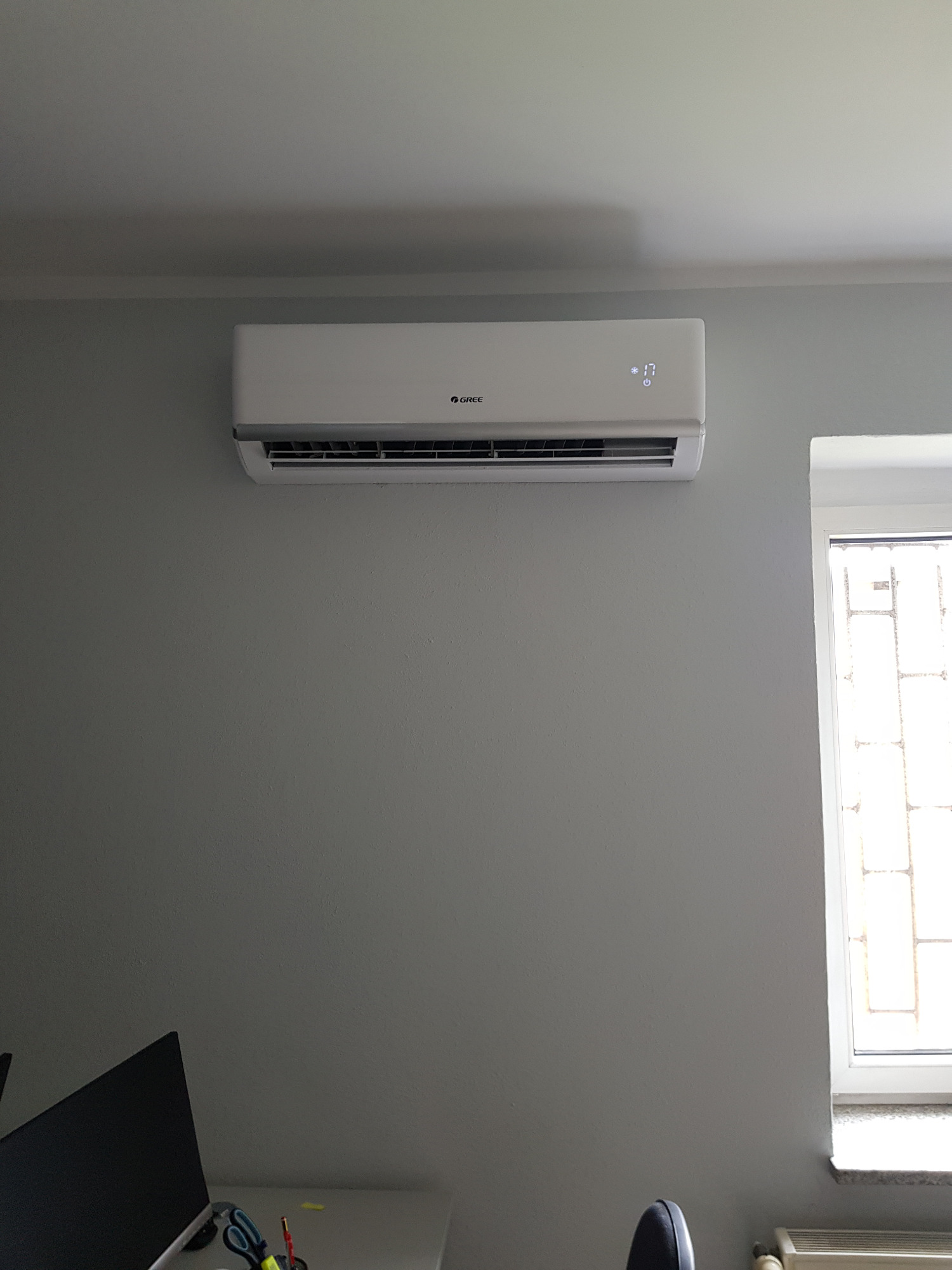 Biały klimatyzator Gree zamontowany na szarej ścianie w biurze, obok okna i nad biurkiem z monitorem.