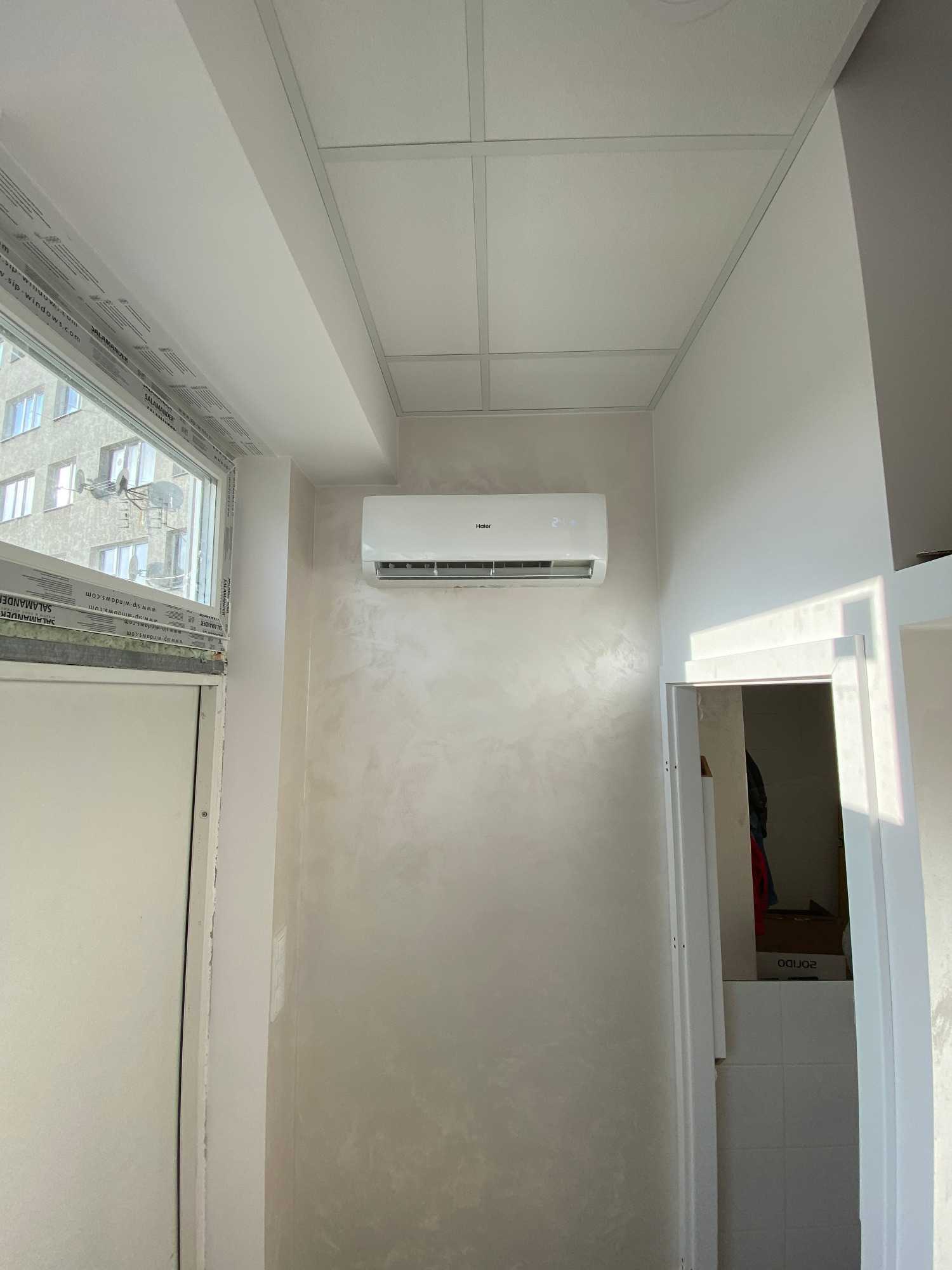 Zamontowany klimatyzator Haier na ścianie z dekoracyjnym tynkiem, widok z dołu, okno z widokiem na budynek mieszkalny, sufit kasetonowy.