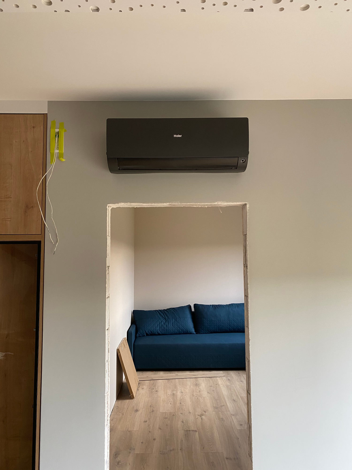 Zamontowana, czarna klimatyzacja marki Haier na szarej ścianie w pokoju, widok z korytarza z widoczną niebieską sofą i drewnianą podłogą.