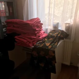 Stos czerwonych tkanin leżący na biurku obok okna z firanką, obok leży wzorzysta tkanina w odcieniach zieleni i brązu.