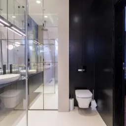 Nowoczesna łazienka z czarnymi szafkami, owalną umywalką nablatową, lustrami w panelach i kabiną prysznicową typu walk-in, widok na sypialnię przez otwarte drzwi.