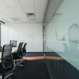 Nowoczesna sala konferencyjna z długim stołem, czarnymi krzesłami na kółkach, przeszkloną ścianą z abstrakcyjnym wzorem i oknami z widokiem na miasto. Na ścianie tablica suchościeralna z notatkami.