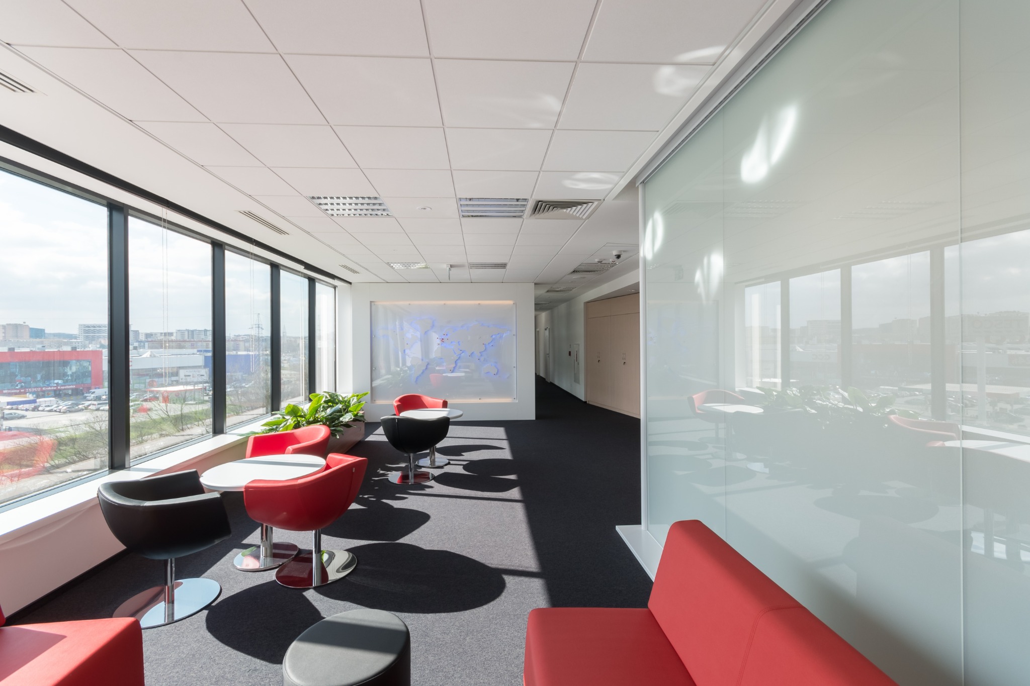 Jasna przestrzeń biurowa z dużymi oknami, czerwonymi i czarnymi krzesłami, białymi stolikami oraz ścianą z mapą świata w tle.