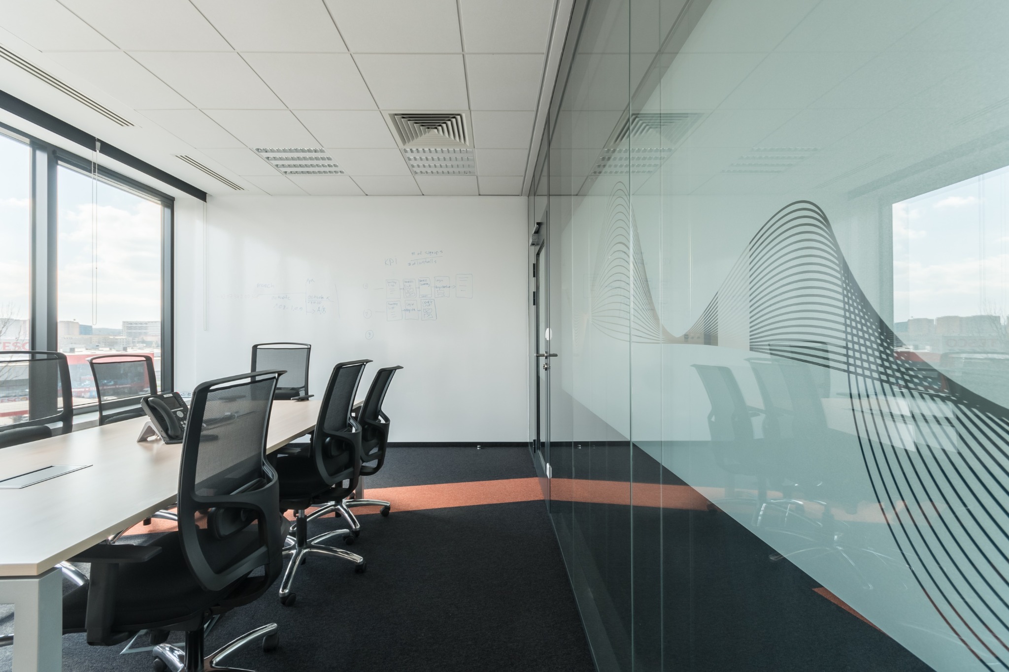 Nowoczesna sala konferencyjna z długim stołem, czarnymi krzesłami na kółkach, przeszkloną ścianą z abstrakcyjnym wzorem i oknami z widokiem na miasto. Na ścianie tablica suchościeralna z notatkami.