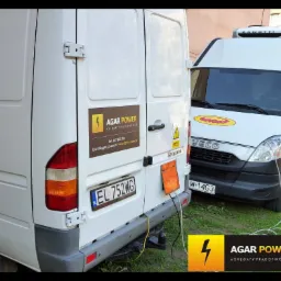 Dwa białe busy zaparkowane obok siebie, z logo firmy Agar Power na tylnych drzwiach jednego z nich, podłączony przewód zasilający do gniazda w busie.