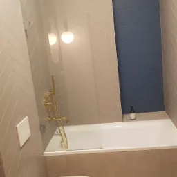 Nowoczesna łazienka z beżowymi płytkami ułożonymi w jodełkę, akcentem w postaci pionowego pasa granatowych płytek strukturalnych, złotą armaturą, białą wanną z parawanem i podwieszaną toaletą.