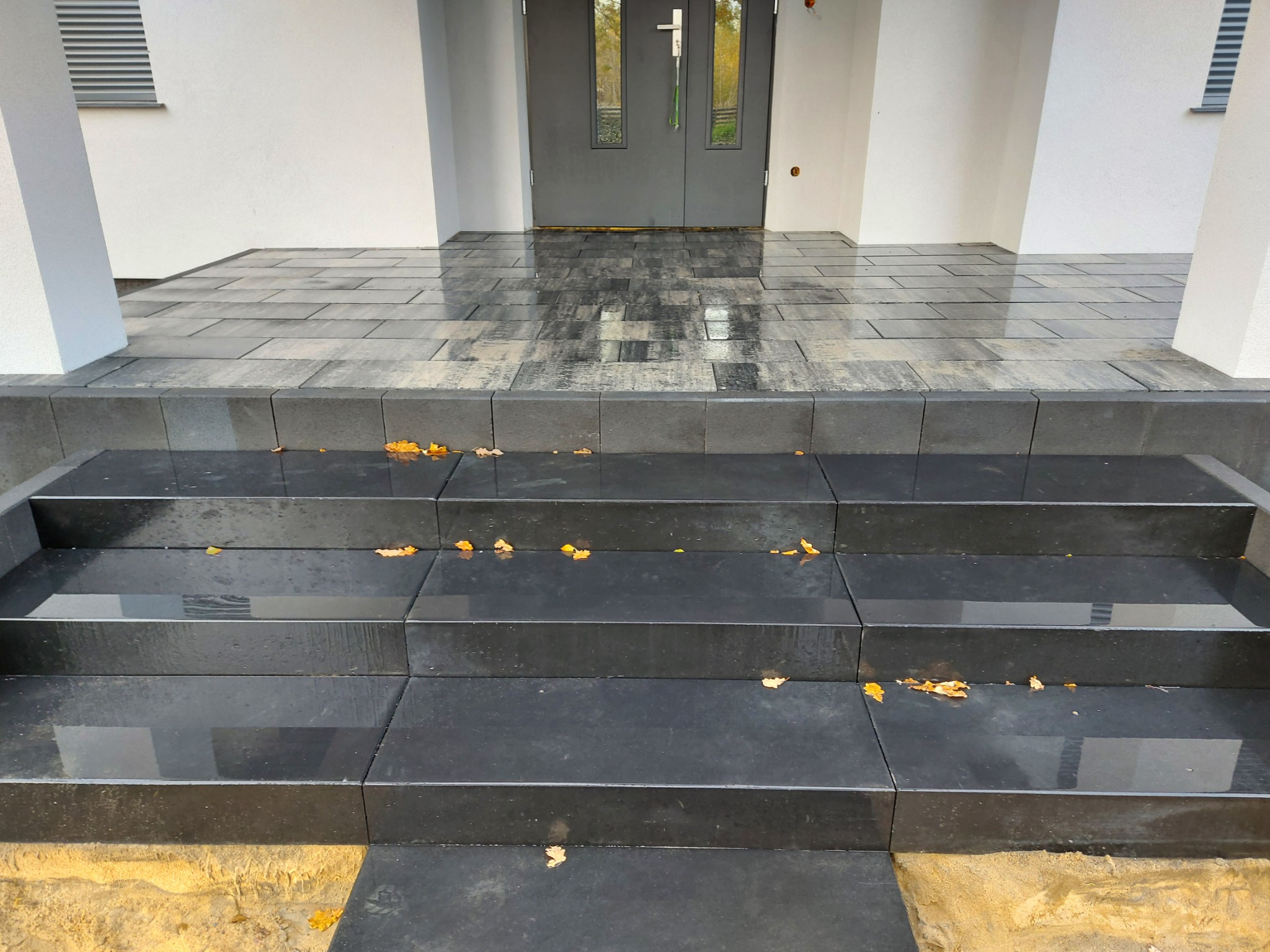 Granitowe schody wejściowe do domu, wykończone czarną kostką, z widocznymi spadłymi liśćmi, prowadzące do szarych drzwi wejściowych i jasnej elewacji.