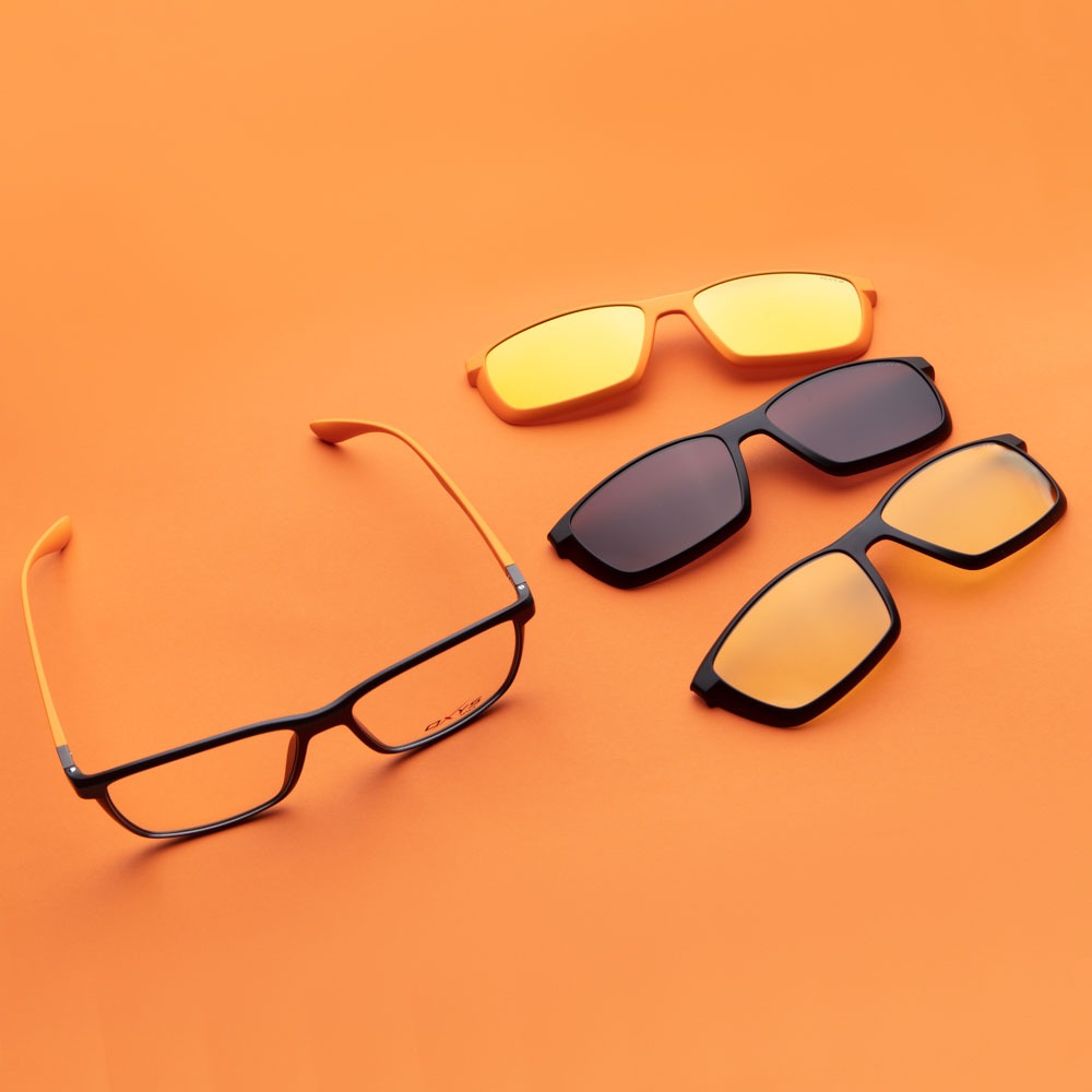 Zestaw okularów korekcyjnych z nakładkami przeciwsłonecznymi w różnych kolorach, ułożonych na pomarańczowym tle. Oprawki czarne z pomarańczowymi zausznikami. Widoczne logo 'OXY' na jednej z oprawek.