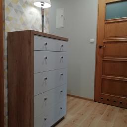 GUD meble - Drewniana komoda z białymi frontami szuflad i okrągłymi, metalowymi uchwytami, stojąca w korytarzu na drewnianej podłodze, obok drewnianych drzwi z szybą i lampy z białym abażurem.