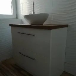 Biała szafka łazienkowa z dwiema szufladami, drewnianym blatem i okrągłą umywalką nablatową, widok z przodu na tle ściany z białych płytek z falistym wzorem.