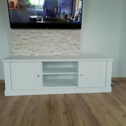 GUD meble - Biała szafka RTV z dwojgiem drzwi i otwartą półką, umieszczona pod telewizorem na ścianie wyłożonej dekoracyjną cegłą, na podłodze z paneli.