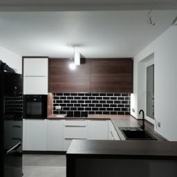 GUD meble - Nowoczesna kuchnia z białymi i drewnianymi szafkami, czarnym blatem i czarnymi płytkami ułożonymi w cegiełkę, oświetlona wiszącą lampą z żarówką, widoczny czarny sprzęt AGD.