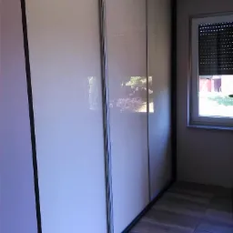 Trzydrzwiowa szafa wnękowa z przesuwnymi, jasnymi frontami, widoczna na tle parkietu i okna z żaluzjami.