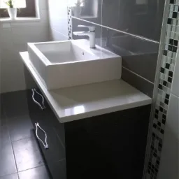 Nowoczesna łazienka z prostokątną umywalką nablatową, czarnym szafką z poziomymi żłobieniami i dekoracyjną mozaiką przy prysznicu.