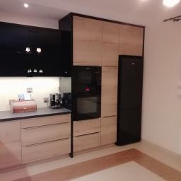 GUD meble - Nowoczesna kuchnia z frontami w kolorze jasnego drewna i czarnymi szafkami górnymi, zabudowana lodówka i piekarnik, widoczne akcesoria kuchenne na blacie.