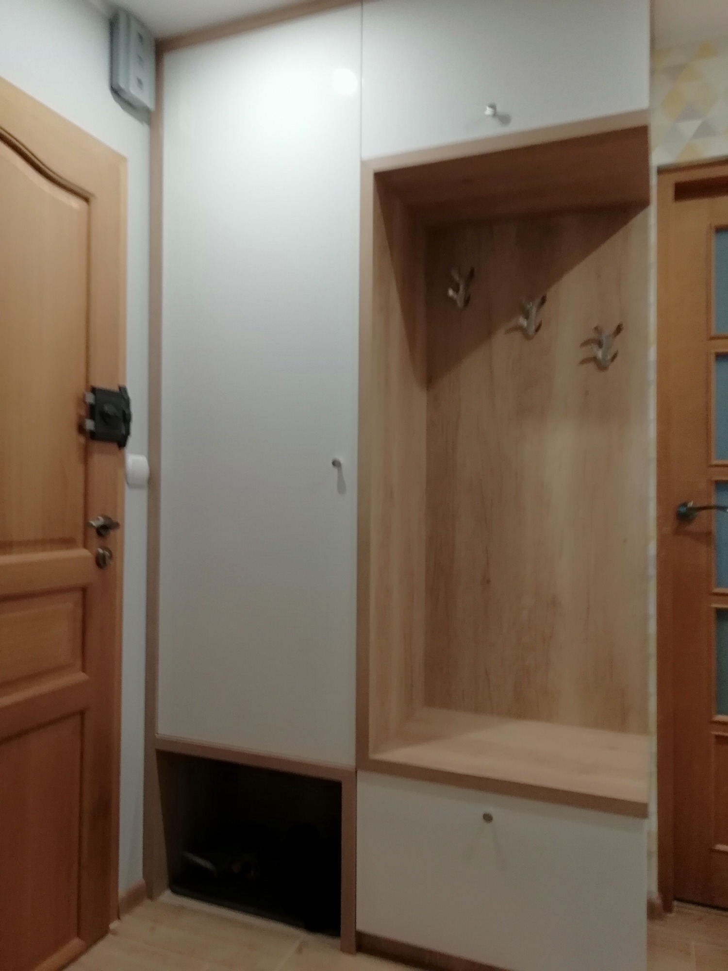 Biało-drewniana szafa wnękowa z otwartą przestrzenią na wieszaki i dolną szafką, widoczna obok drewnianych drzwi wejściowych.