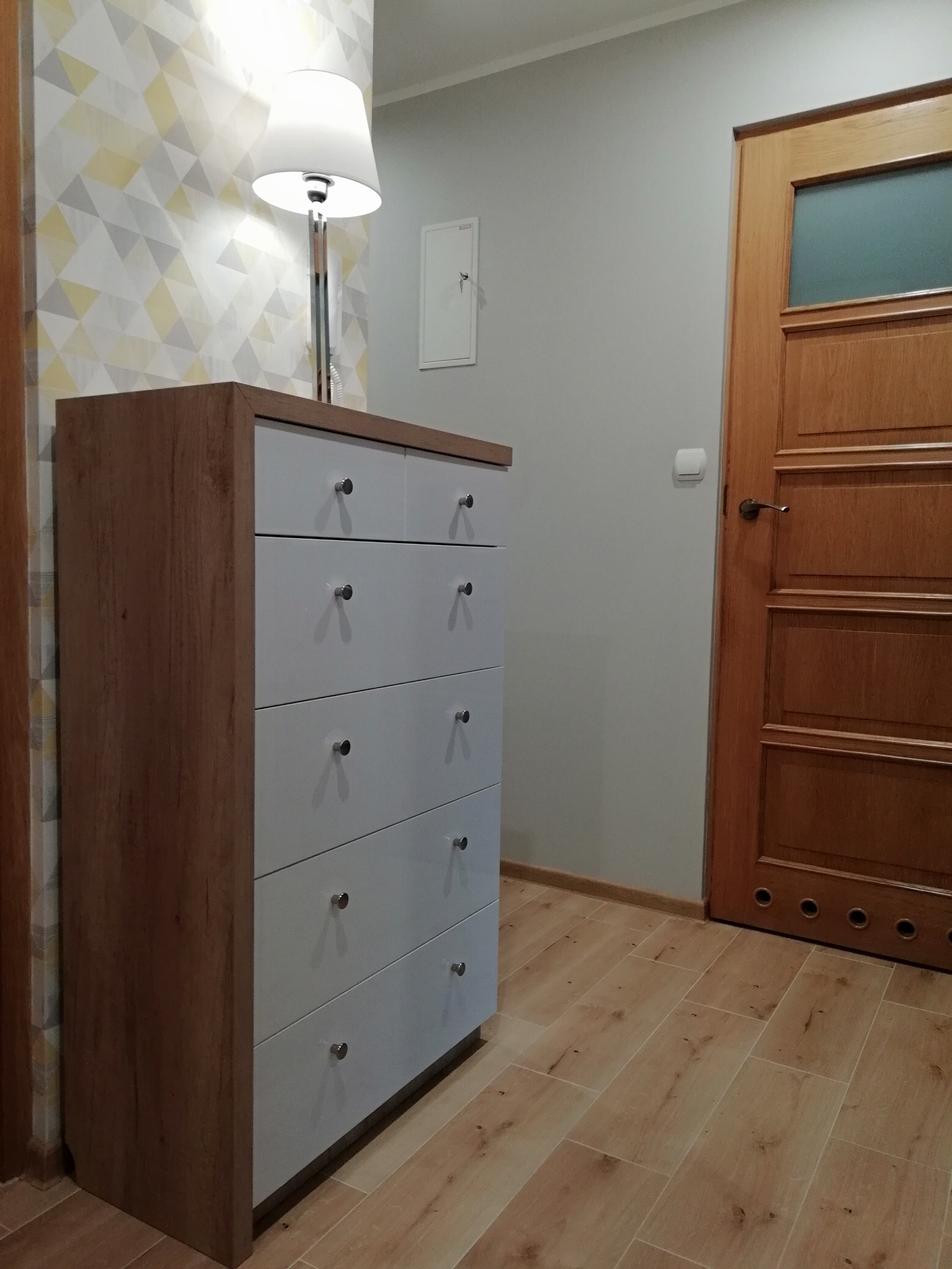 Drewniana komoda z białymi frontami szuflad i okrągłymi, metalowymi uchwytami, stojąca w korytarzu na drewnianej podłodze, obok drewnianych drzwi z szybą i lampy z białym abażurem.