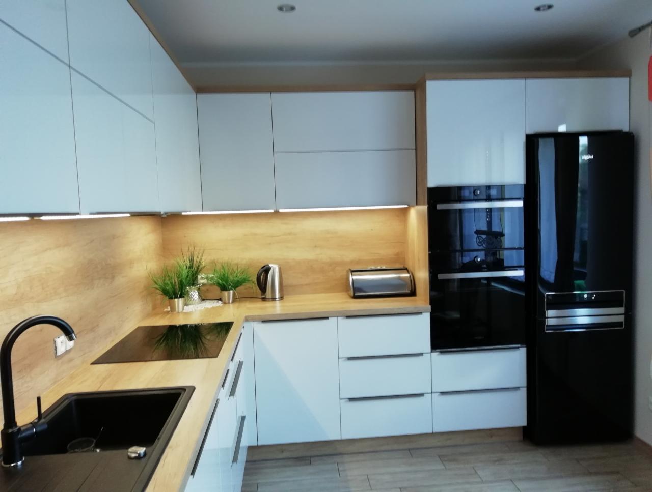 Nowoczesna kuchnia z białymi, gładkimi frontami szafek, drewnianym blatem i czarnym zlewem oraz sprzętem AGD w kolorze czarnym, oświetlona ciepłym światłem LED pod szafkami.