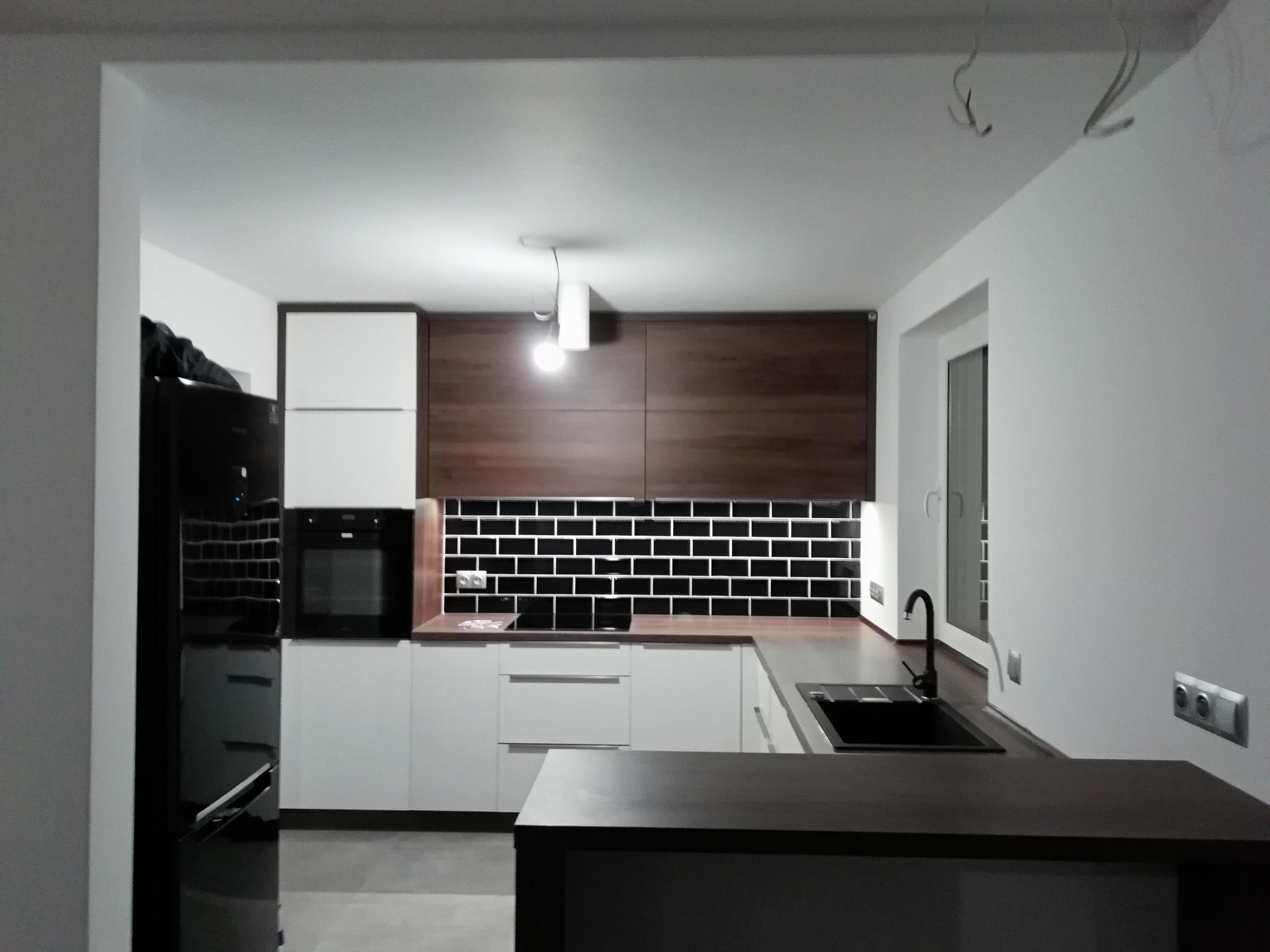 Nowoczesna kuchnia z białymi i drewnianymi szafkami, czarnym blatem i czarnymi płytkami ułożonymi w cegiełkę, oświetlona wiszącą lampą z żarówką, widoczny czarny sprzęt AGD.