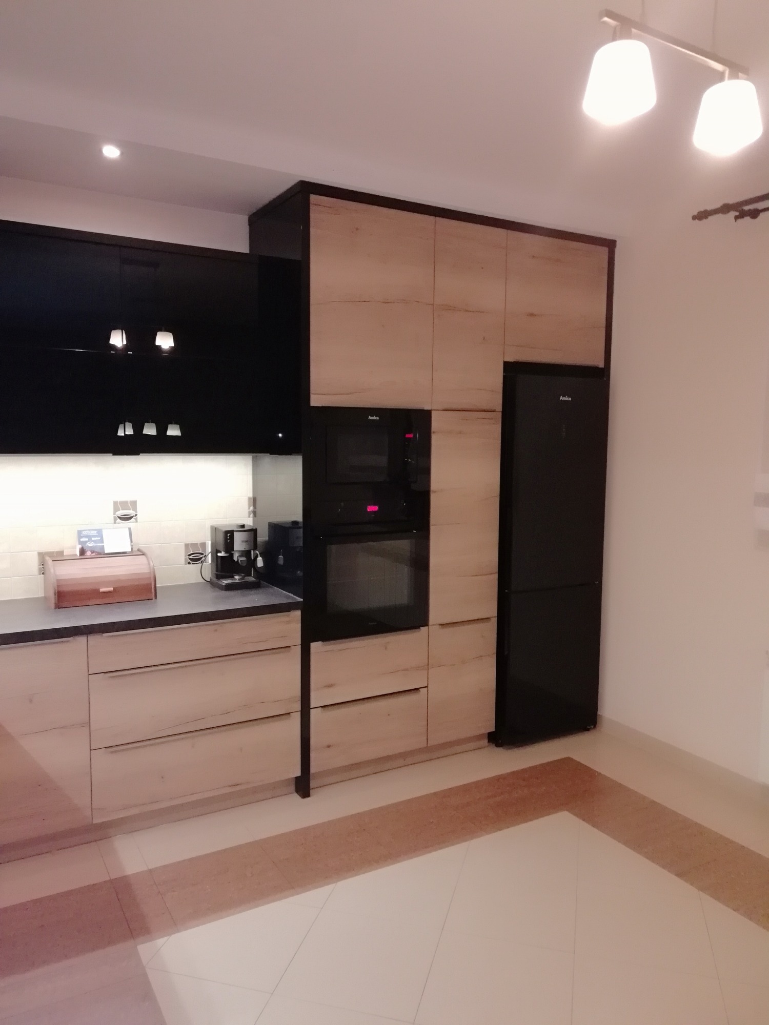 Nowoczesna kuchnia z frontami w kolorze jasnego drewna i czarnymi szafkami górnymi, zabudowana lodówka i piekarnik, widoczne akcesoria kuchenne na blacie.