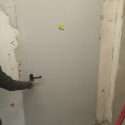 P.W.KOWAL Daniel Kowalski - Montaż szarych drzwi wejściowych z czarną klamką w surowym otoczeniu, widoczne ślady po remoncie na ścianach.