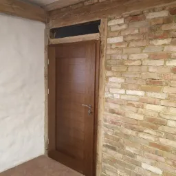 Nowe, brązowe drzwi wewnętrzne z prostą klamką, osadzone w surowym otworze z odsłoniętą cegłą i drewnianymi belkami stropowymi, na podłodze terakota.