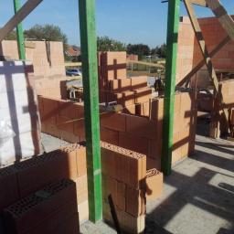 P.W.KOWAL Daniel Kowalski - Wznoszenie ścian zewnętrznych z czerwonej cegły ceramicznej, widoczne zielone stemple budowlane i drewniane podpory stabilizujące konstrukcję, etap budowy domu jednorodzinnego.