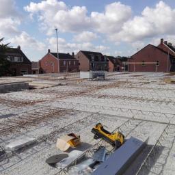 P.W.KOWAL Daniel Kowalski - Zbrojenie płyty fundamentowej z widocznymi elementami szalunku i narzędziami budowlanymi na tle zabudowy jednorodzinnej pod błękitnym niebem.