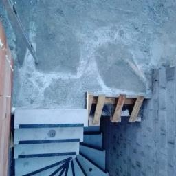 P.W.KOWAL Daniel Kowalski - Wewnętrzne schody spiralne z szarych płyt z czarnymi akcentami podczas budowy, widoczne drewniane podpory i surowy beton na ścianach.