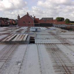 P.W.KOWAL Daniel Kowalski - Zbrojenie stropu w trakcie budowy domu jednorodzinnego, widoczne drewniane deski szalunkowe i stalowe pręty zbrojeniowe, w tle zabudowa miejska i pochmurne niebo.