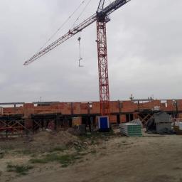 P.W.KOWAL Daniel Kowalski - Budowa domu murowanego w trakcie realizacji, widoczny fragment konstrukcji z cegły, dźwig budowlany i materiały budowlane na placu budowy w pochmurny dzień.